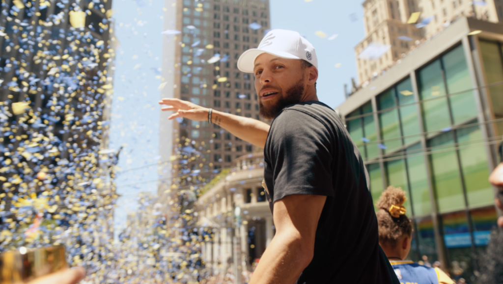 Stephen Curry at Warriors parade. Confetti falling, white cap, black shirt. NBA champion celebration.