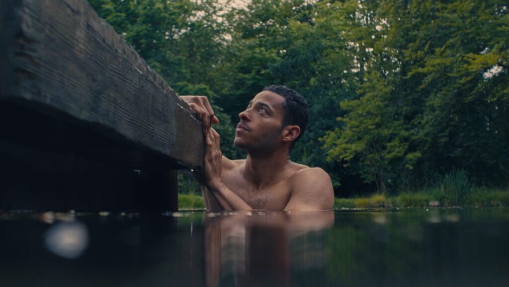 Shirtless man in water, grasping wooden dock, looking up. Nature scene with trees. The Lesson movie still.