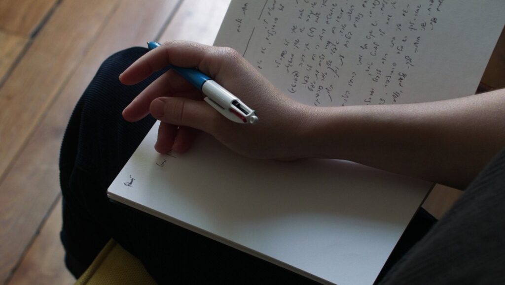 Hand holding pen, writing on paper. The Night Drags On film review. Mathilde Girard, FIDMarseille '23.