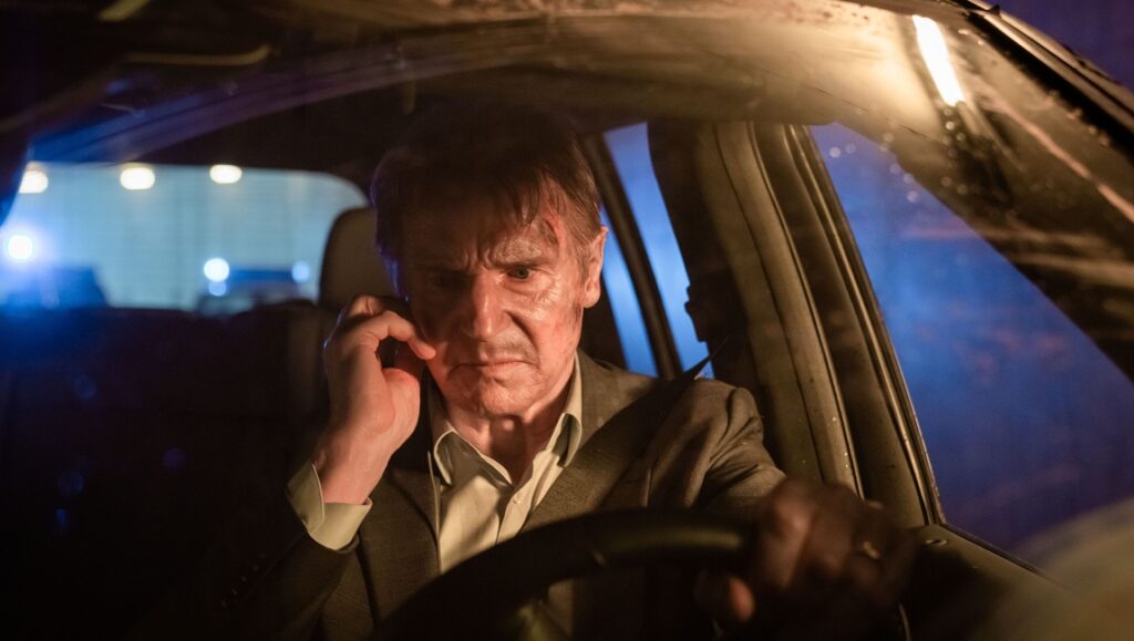 Liam Neeson in Retribution. Actor driving a car, looking stressed and holding a phone to his ear.