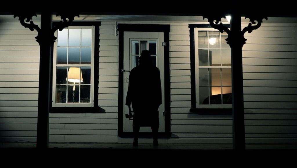 Movie scene from Where the Devil Roams. Dark figure with an axe stands before a house. Horror film concept.