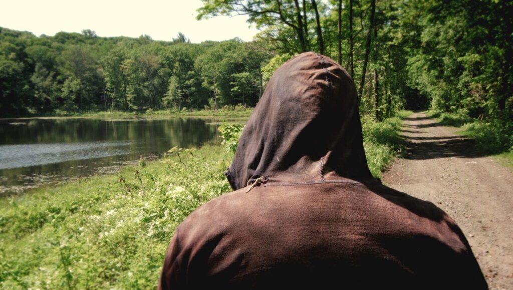 Brightwood — Dane Elcar Brightwood still: Person in hood by lake. Dane Elcar film scene. Forest path.