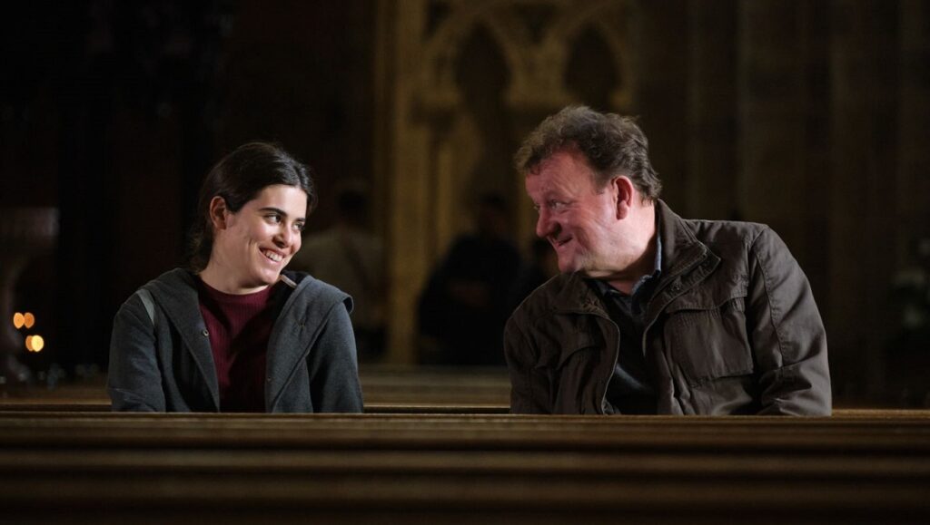 Dreaming & Dying film still. Woman and man smile in church pews. Locarno '23 review.