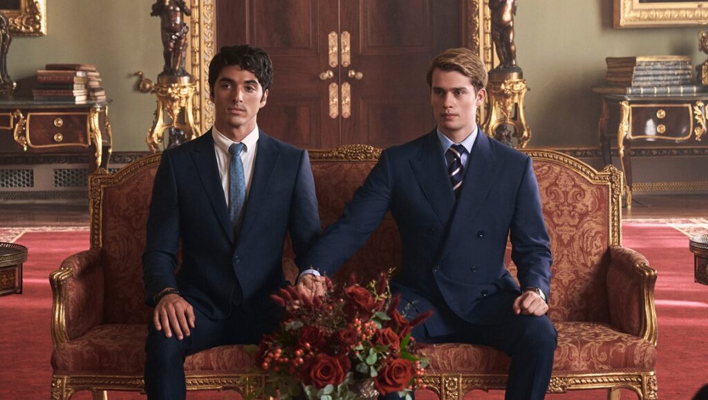 Red, White & Royal Blue movie still. Two men in suits sit together holding hands.