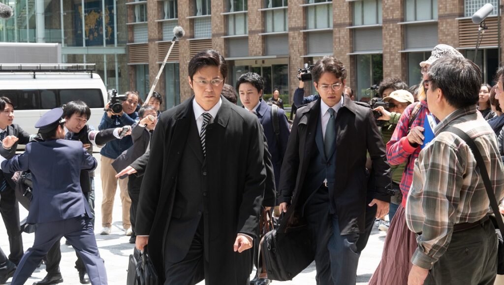 Japanese drama scene. Men in suits walk through a crowd with cameras and microphones.