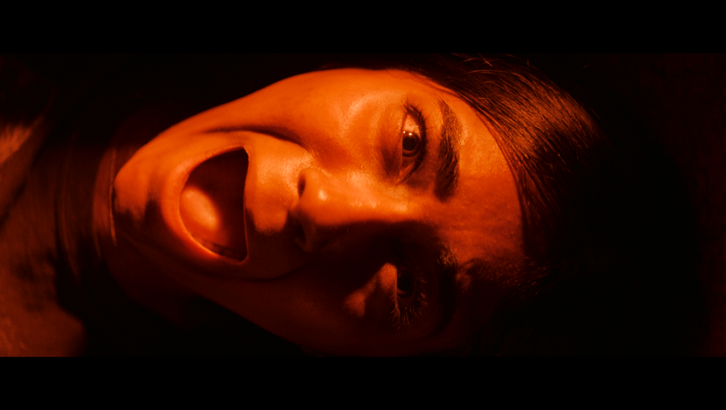 It Lives Inside — Bishal Dutta Close-up of terrified Samidha, from It Lives Inside, mouth open in a scream, bathed in orange light.