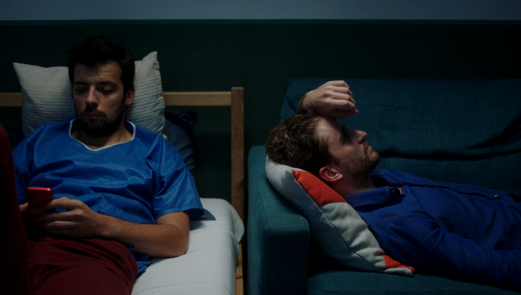 MMXX film scene: Two men in blue shirts, one using a phone, the other reclining on a couch. Cristi Puiu SSSIFF '23 review.