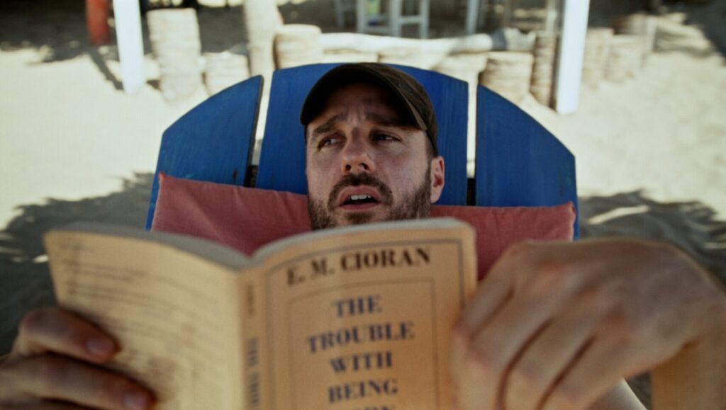 Sebastian Silva in Rotting in the Sun movie. Man reading The Trouble With Being Born on beach.