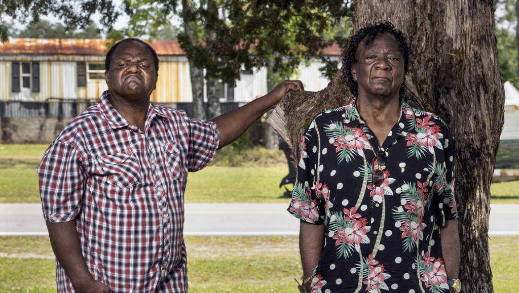 Silver Dollar Road image. Two men stand near a tree. One wears a plaid shirt, the other a floral shirt.