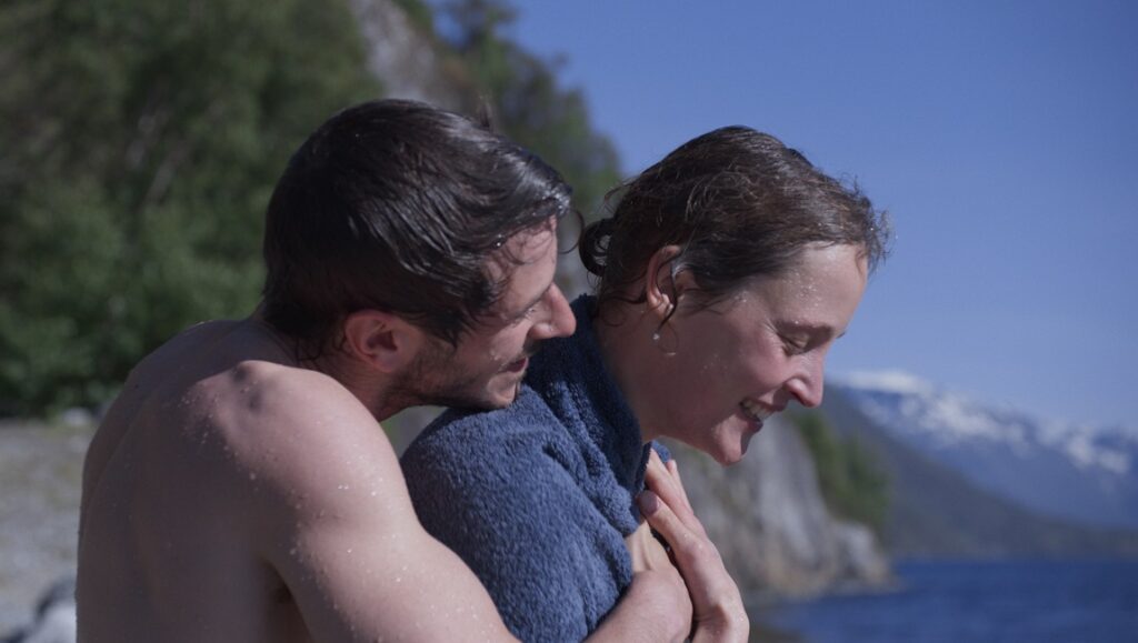 More Than Ever — Emily Atef More Than Ever film still: A man embraces a woman with a towel. Mountains and water in the background.