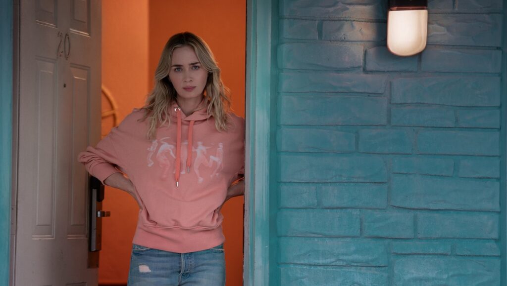 Emily Blunt in Pain Hustlers. Blunt wears a pink hoodie and jeans, standing in a doorway with a concerned expression.