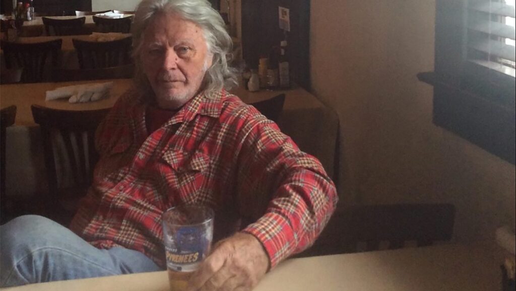 James Benning interview. Portrait of the filmmaker in a red flannel shirt, holding a glass of beer.