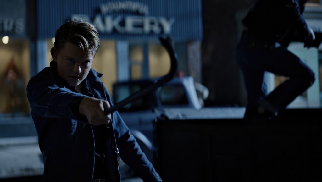 Dark Harvest movie still. Teen boy holds weapon in front of bakery at night.