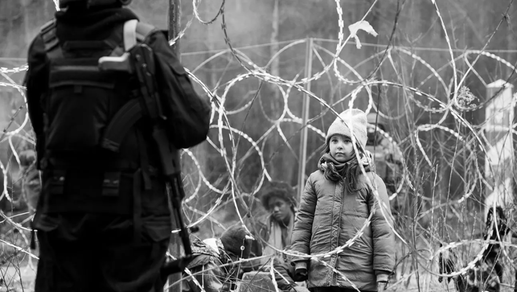 Green Border film still. Child refugee behind barbed wire, guarded by armed soldier. Agnieszka Holland film review.
