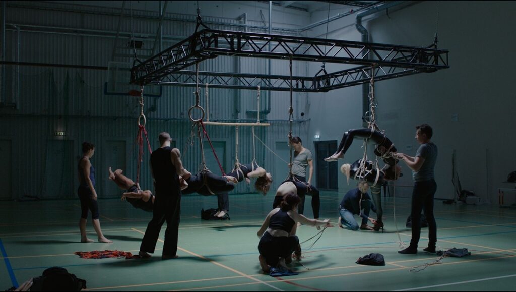 Dark Paradise film scene. People suspended by ropes in a gym-like setting. Tallinn Black Nights Film Festival review.