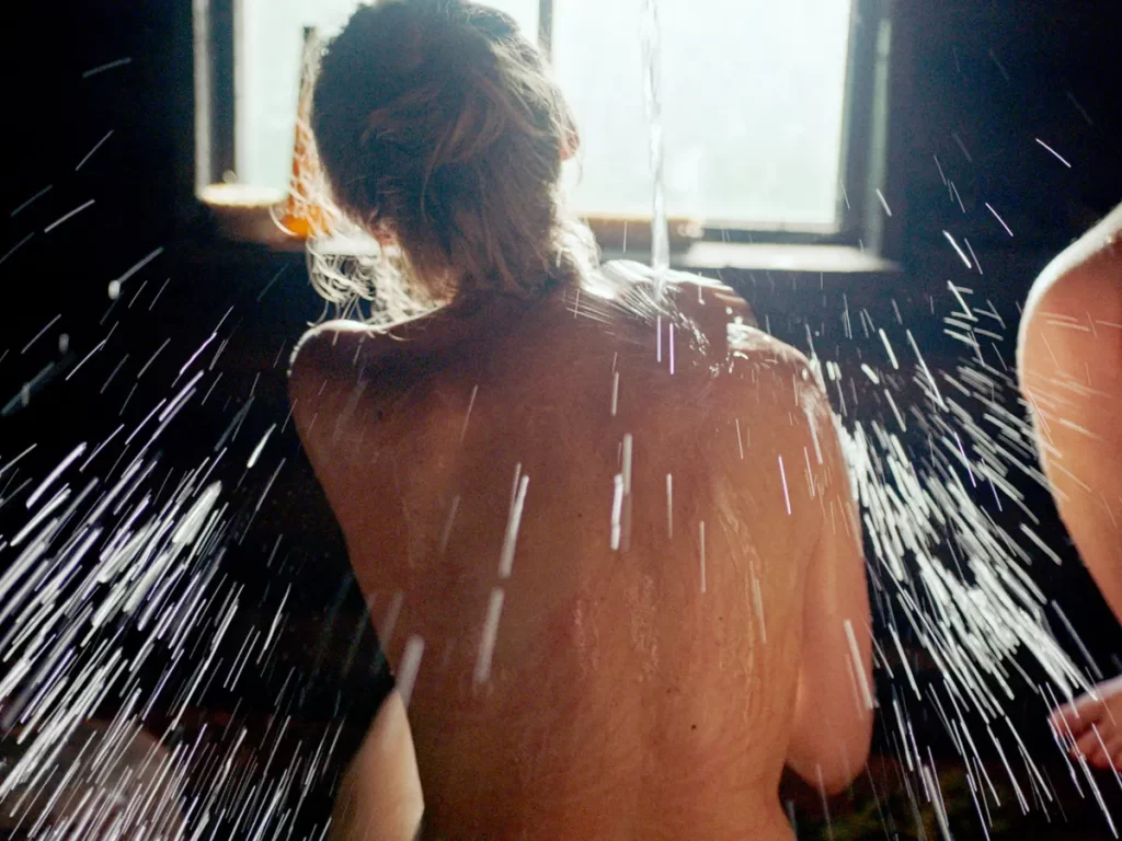 Woman in smoke sauna. Water pouring on back, window in background. Smoke Sauna Sisterhood.