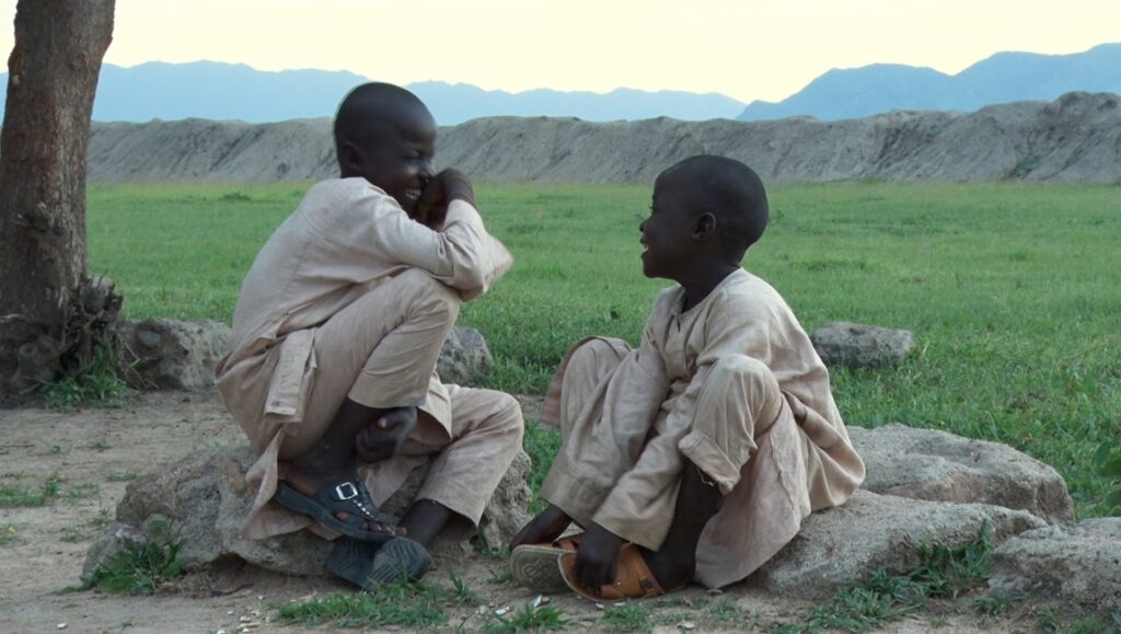 Two boys in They Shot the Piano Player, sitting on rocks in a field, chatting. Mountains in background.