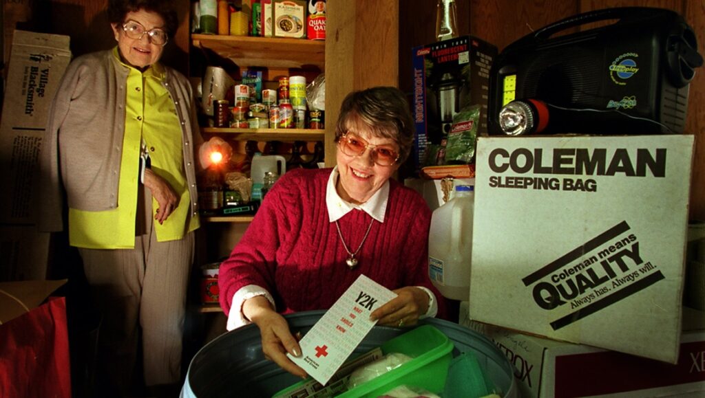 Time Bomb Y2K — Marley McDonald and Brian Becker They Shot the Piano Player Y2K panic scene. Two women with a Y2K info sheet, surrounded by supplies and a Coleman sleeping bag.