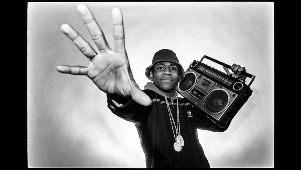 Black and white portrait of a rapper with a boombox, wearing a bucket hat and gold chains.