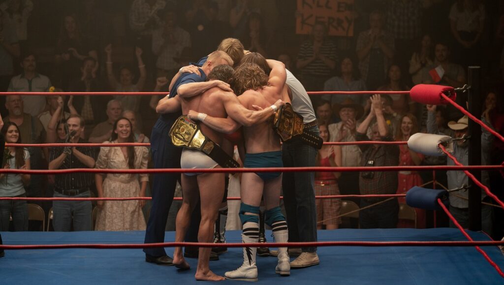 The Iron Claw — Sean Durkin The Iron Claw movie still: Von Erich wrestling family embrace in the ring, championship belts visible. Crowd cheers.