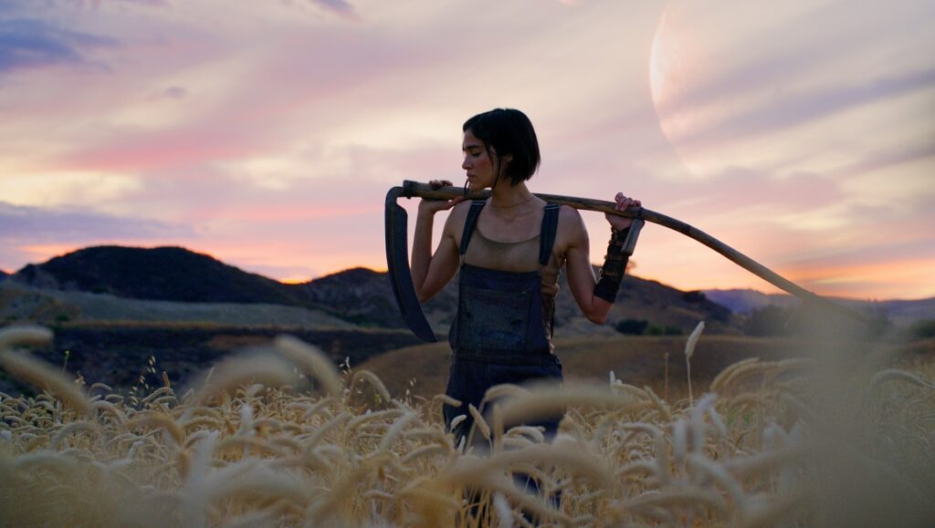 Sofia Boutella as Kora in Zack Snyder's Rebel Moon. Woman with weapon in field at sunset. Sci-fi, adventure movie.