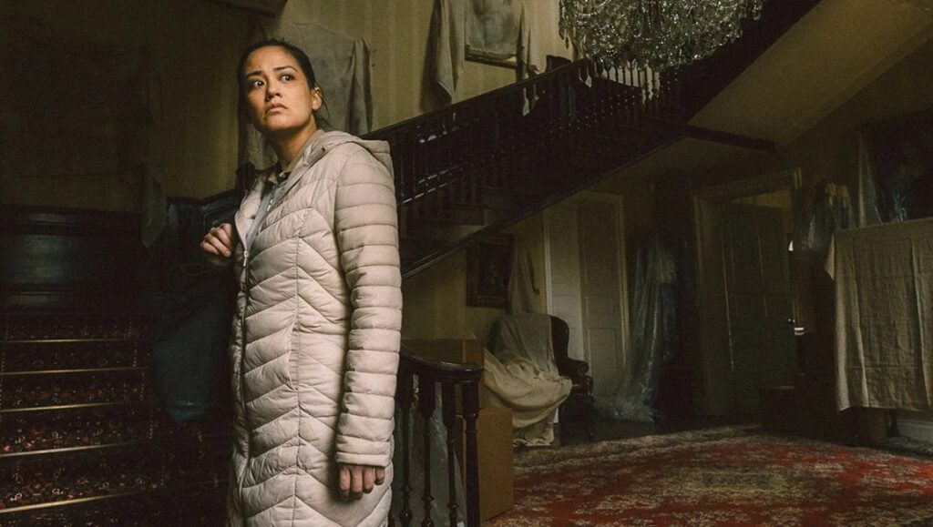 Raging Grace film still. Woman in coat stands in a dark, covered mansion. Staircase and chandelier visible.