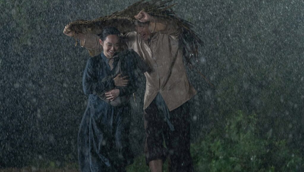The Last Wife — Victor Vu The Last Wife movie scene: A couple huddles in the rain, sheltered by a makeshift cover. Victor Vu film.