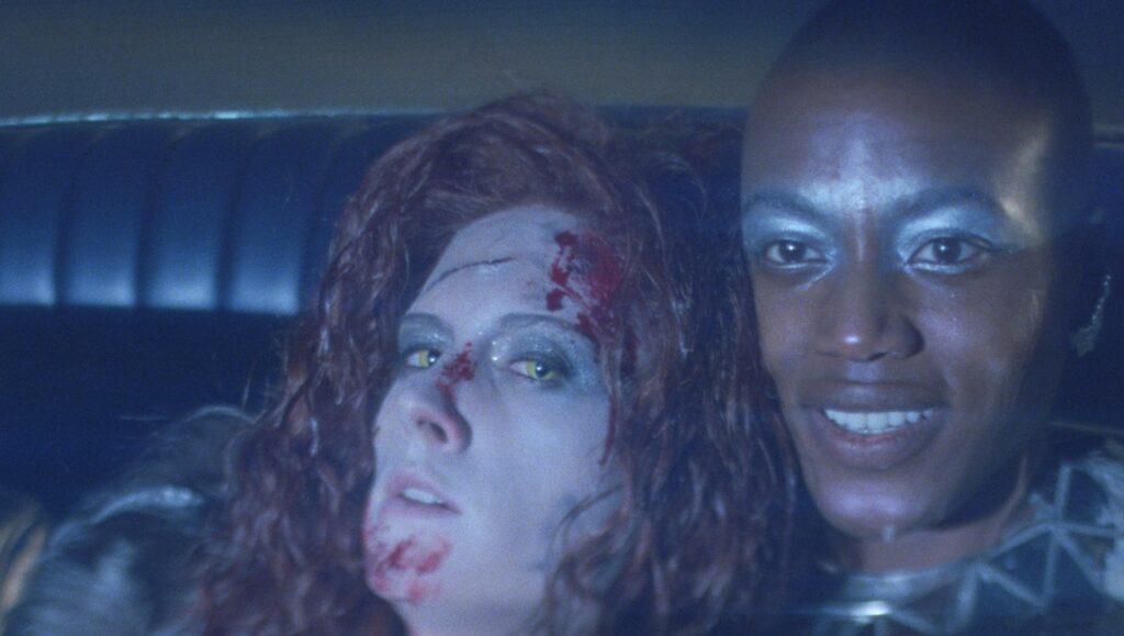 She is Conann film still. Sanja and a bald woman in a car. Blood, yellow eyes, silver makeup.