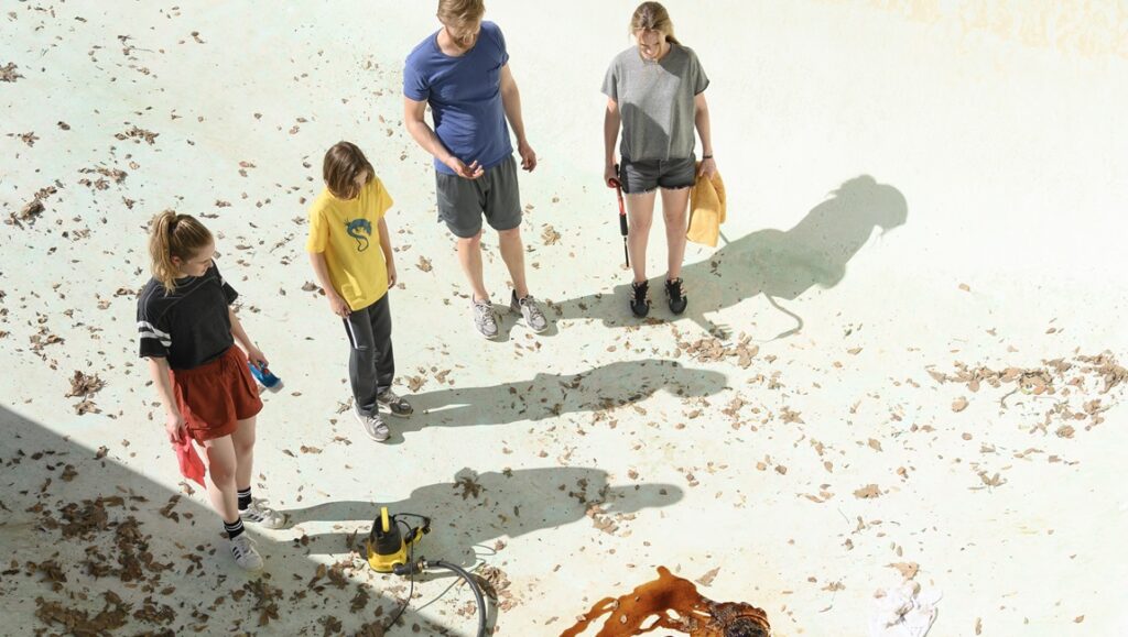 Night Swim movie scene: Family cleaning a dirty, leaf-filled swimming pool. Horror film vibes.
