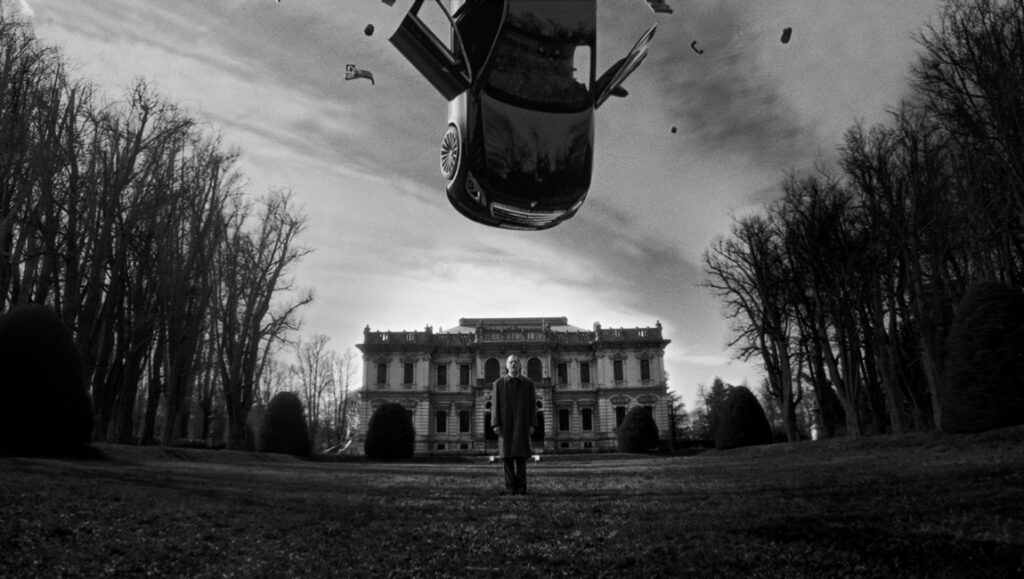 Black and white surreal photo. Man stands before mansion as car falls from sky. Fabio D' Orta art.