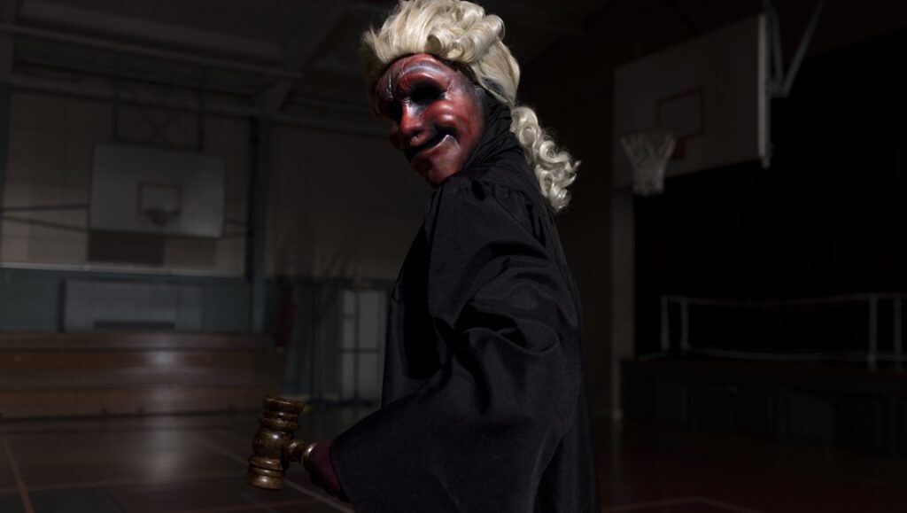 Founders Day movie still. Figure in judge's robe and mask holds gavel in a gymnasium.