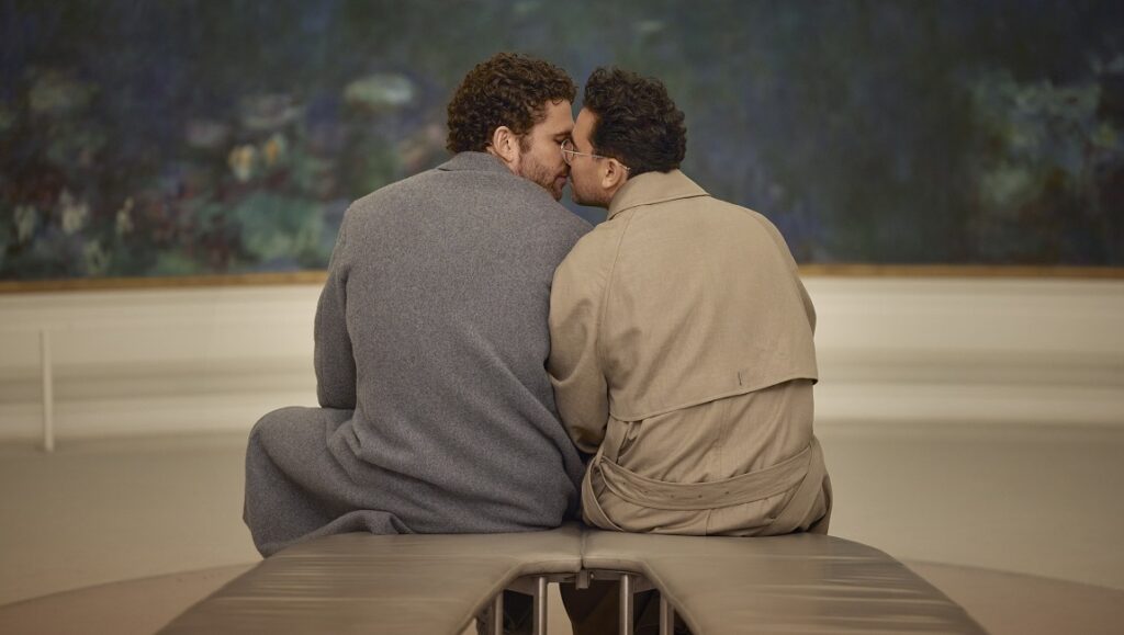 Good Grief movie scene: Dan Levy and a man share a kiss in front of a painting. Romance and grief intertwine.