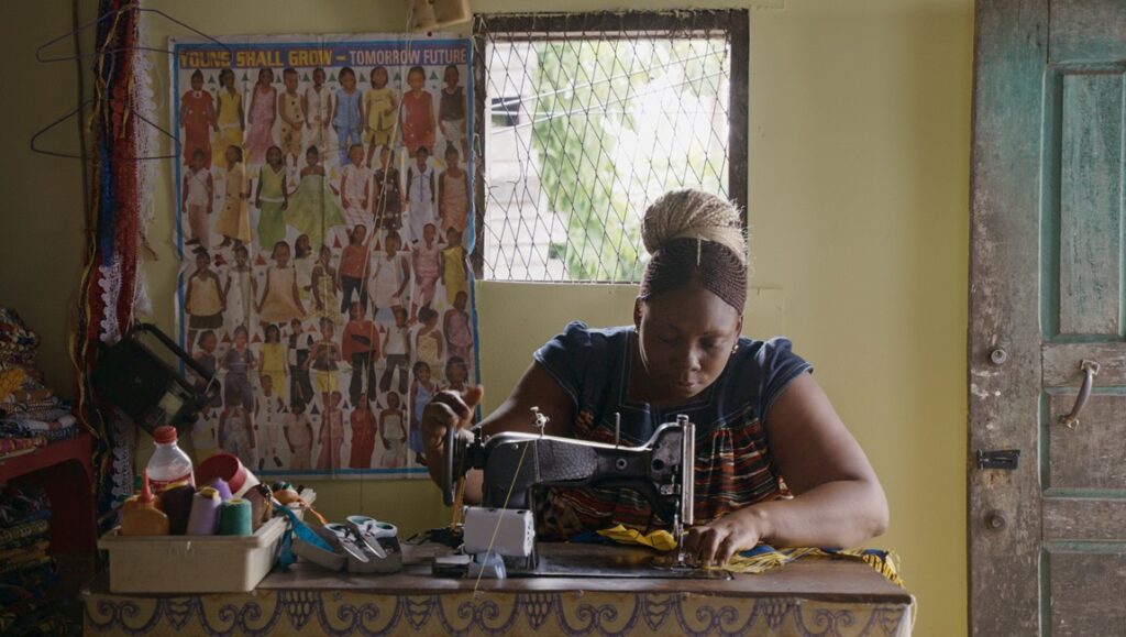Mambar Pierrette, Rosine Mbakam film: Woman sewing with a vintage machine in a small room, window, and poster on the wall.