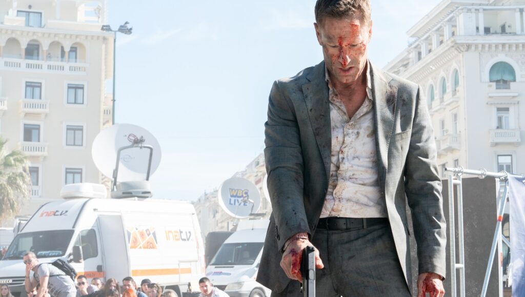 Aaron Eckhart in The Bricklayer movie. Bloodied man in suit holds gun.