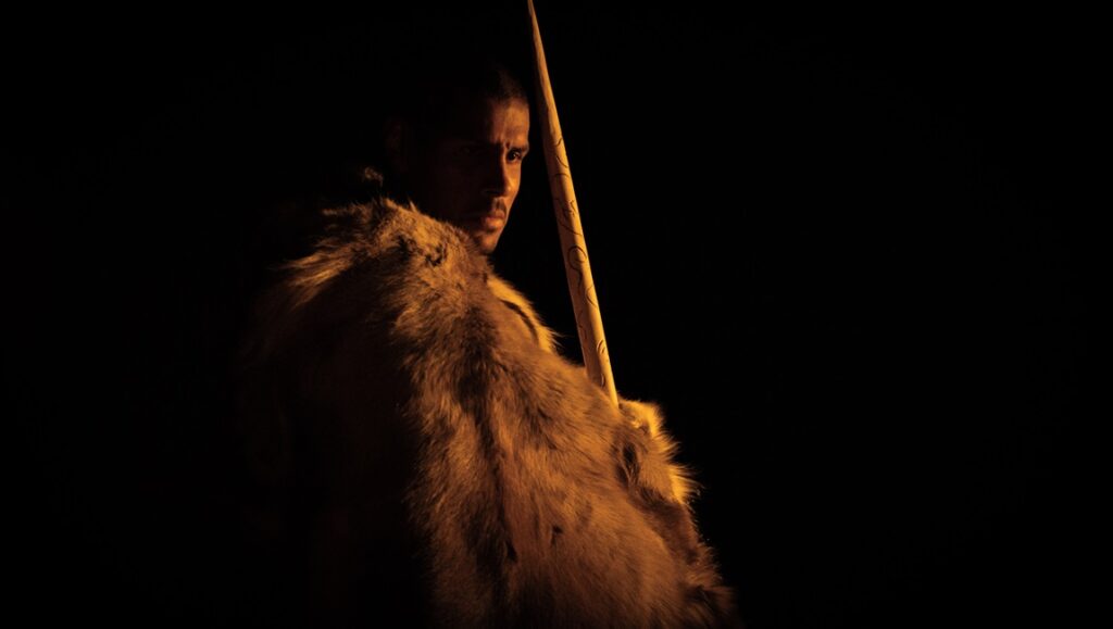 Out of Darkness film still: A man in furs holds a spear, looking back in the dark.
