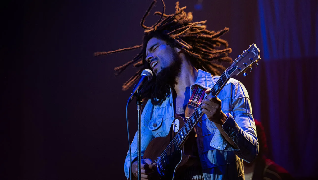 Kinsgley Ben-Adir as Bob Marley in One Love movie. Musician sings into microphone while playing guitar. Dreadlocks fly.