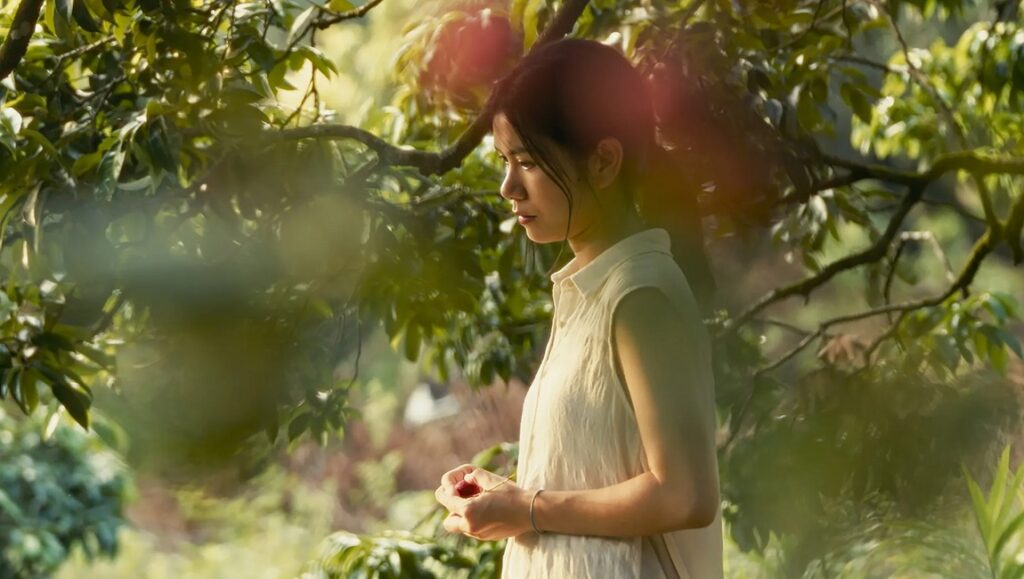 Choy Ji in Borrowed Time (IFFR '24). Woman in nature, holding a red fruit. Film review image.