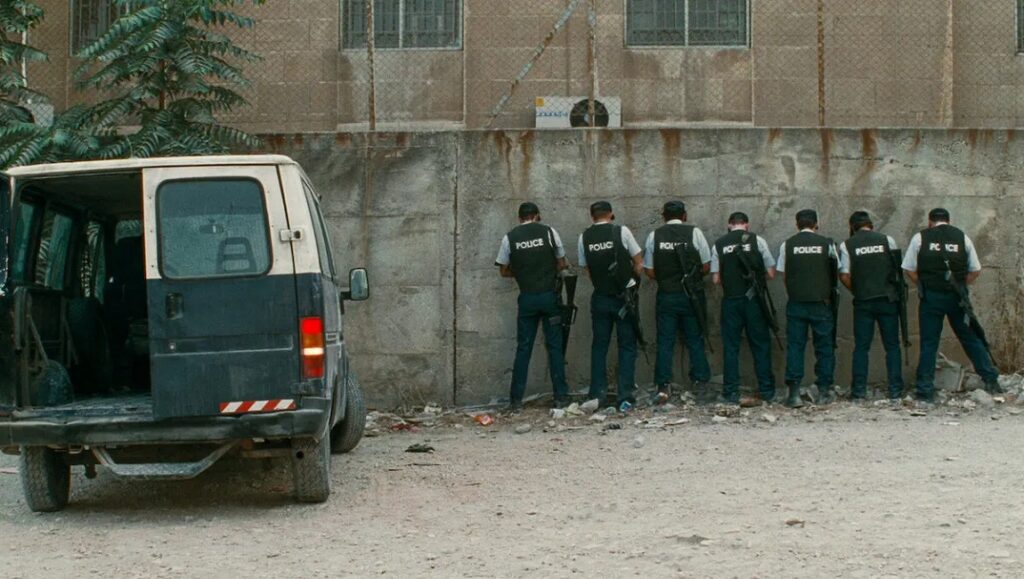 Chronicle of a Disappearance still: Police officers urinating against a wall near a van.