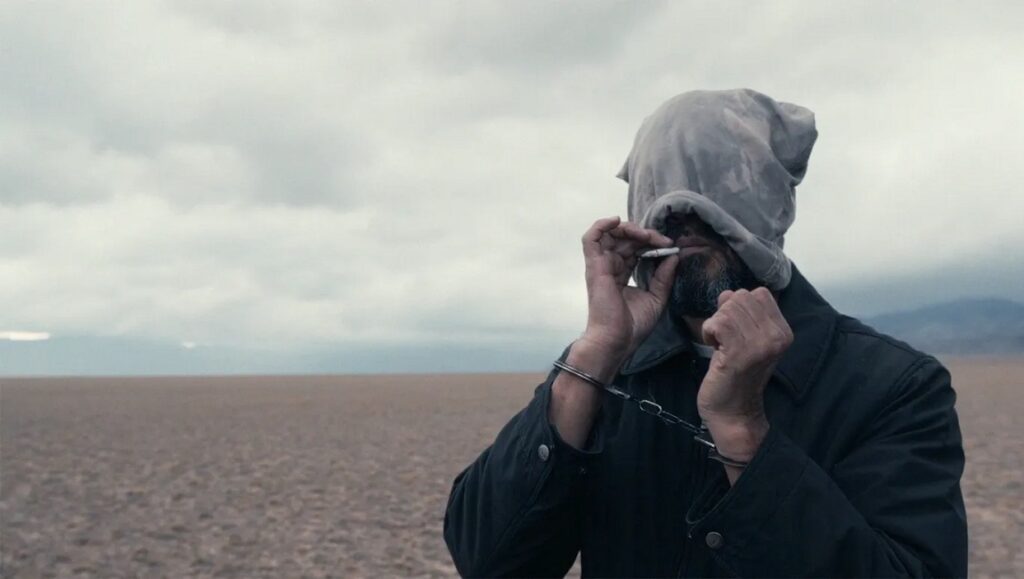 IFFR 2024 — Dispatch 3: Steppenwolf, Schirkoa: In Lies We Trust, Cosmic Miniatures IFFR 2024: Steppenwolf film still. Handcuffed figure with head covering smokes in a desolate landscape. Drama, independent film.
