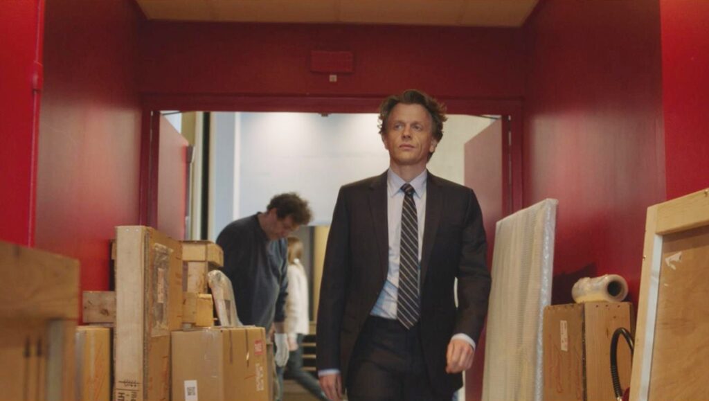 The Book of Solutions film still. Man in suit walks past boxes. Rendez-Vous with French Cinema 2024.