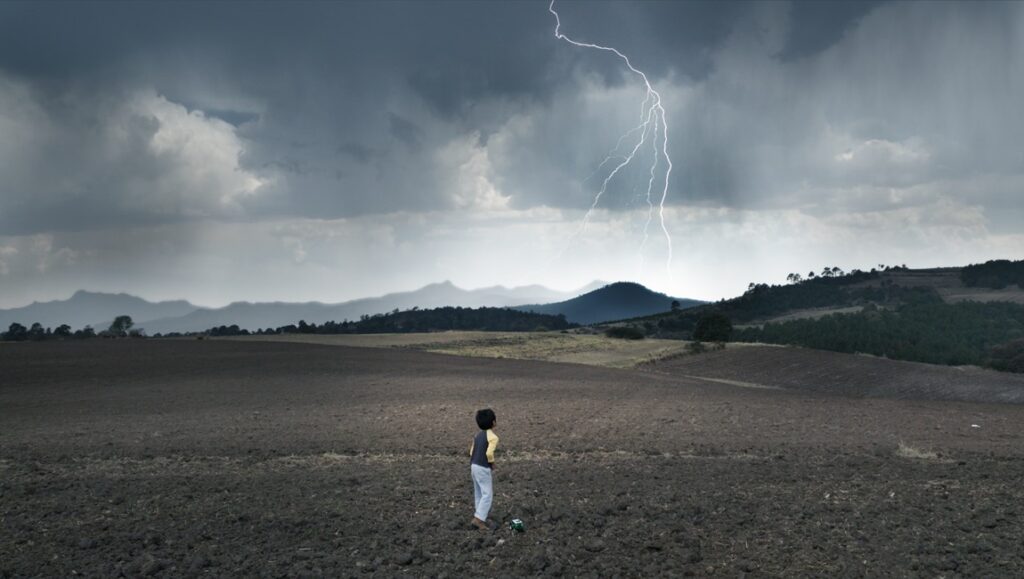 El Eco film still. Child stands in a field as lightning strikes during a storm. Landscape with mountains in the background.