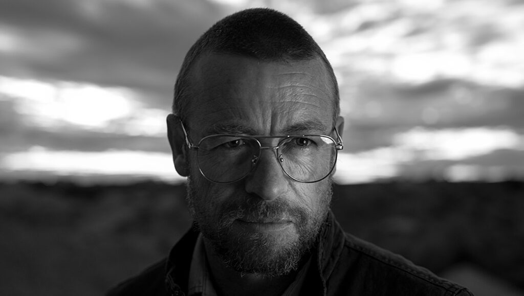 Still from Limbo (Ivan Sen film): Man with glasses in black and white, against a cloudy sky.