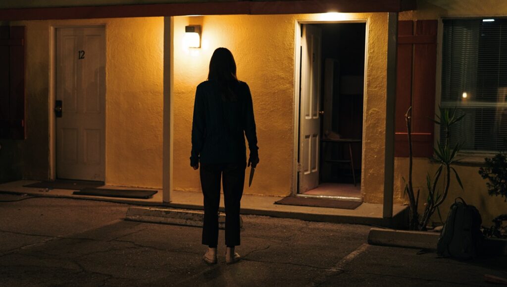 Night Shift movie still. Woman with knife stands outside motel room at night. Horror film scene.