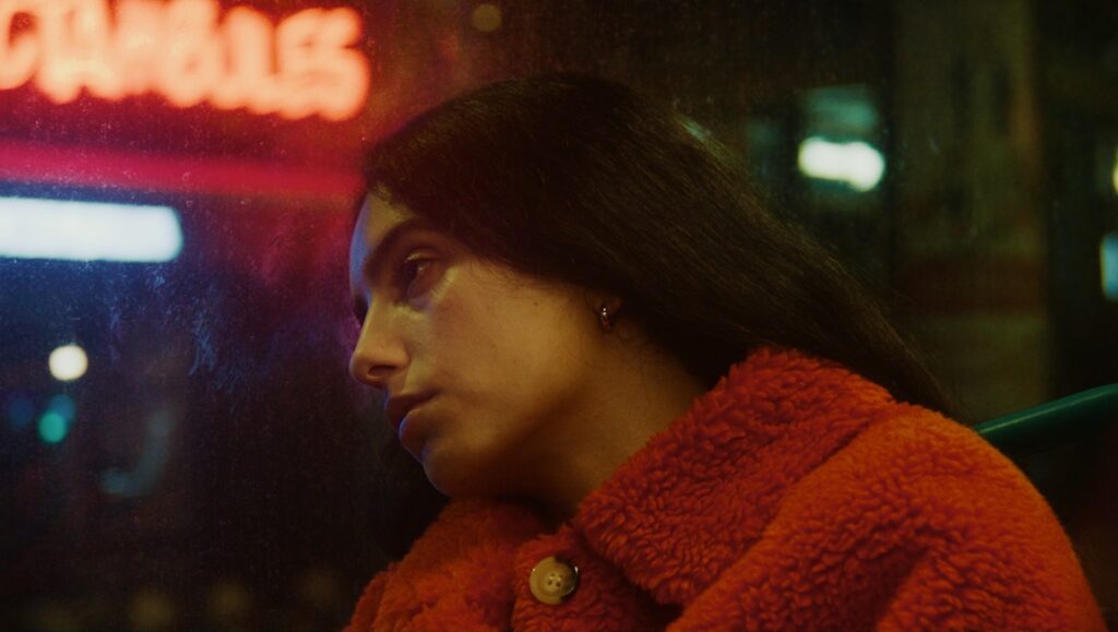 Pensive woman in red coat, city lights. Portrait of a young woman looking out a window at night. Neon sign reflection.