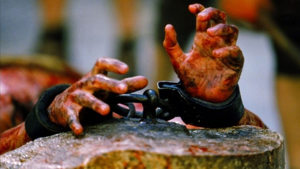 The Passion of the Christ: Bloodied hands bound in shackles on a wooden block.