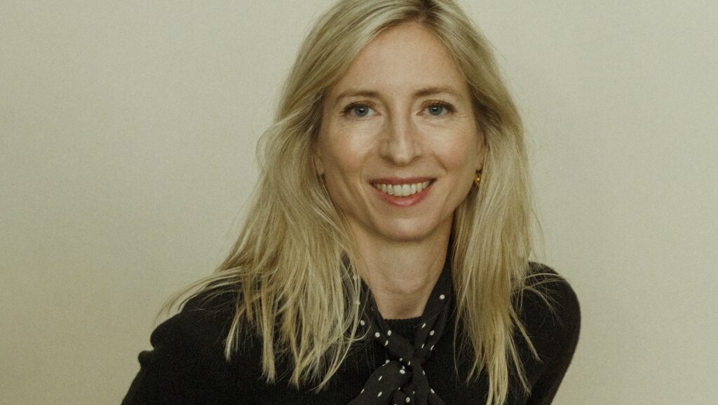 Jessica Hausner portrait. The Austrian film director with blonde hair smiles in front of a neutral background.