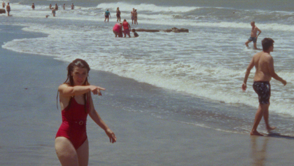 Film still from Republic review. Woman in red swimsuit points at beach with ocean waves and people.