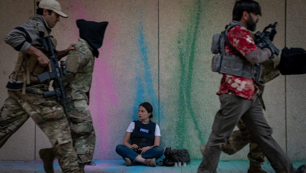 Civil War movie scene: Journalist sits as armed soldiers pass. Review of Alex Garland's film.