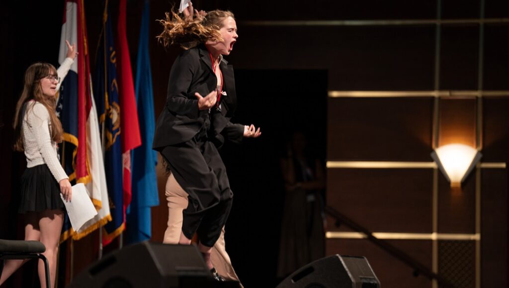 Girls State review: Teen girl in suit jumps on stage, yelling. Flags behind her. Documentary film by Jesse Moss & Amanda McBaine.