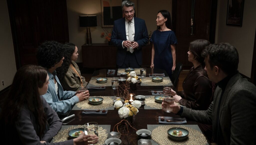 Still from Humane movie review. A family sits at a formal dinner table with a man and woman standing at the head.